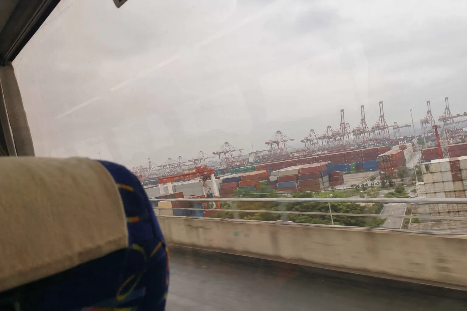 A container port shot through the window of a moving coach: stacked containers and gantry cranes to the horizon, the window frame and a smear of reflection across one corner, a headrest in the foreground. Overcast, flat, slightly motion-blurred.