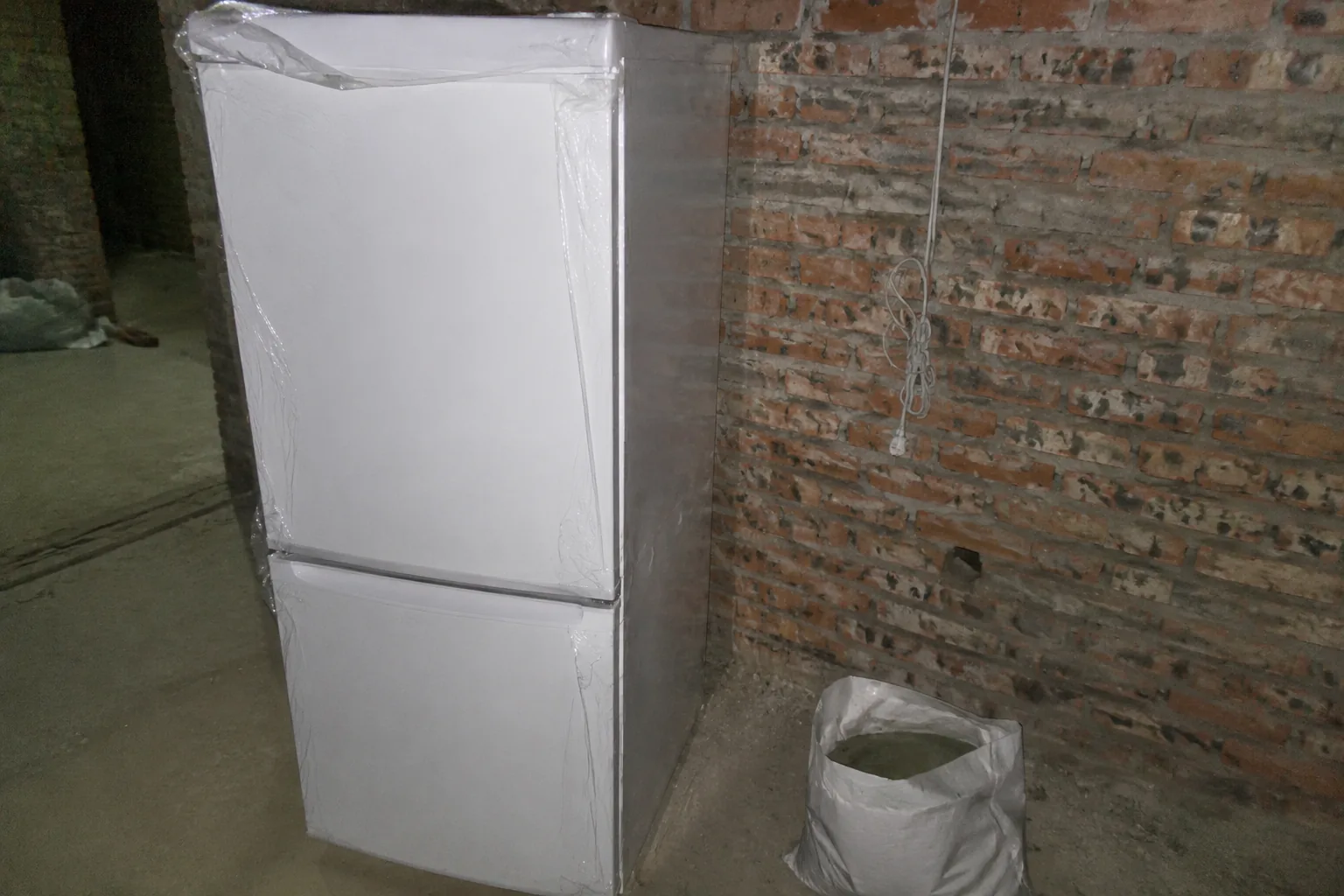 Flash photo inside a half-finished rural house: a brand-new white refrigerator with the protective plastic still on the door, standing on bare concrete against an unplastered brick wall, an exposed cable and a bag of tile adhesive beside it. Blown highlights, dark corners.