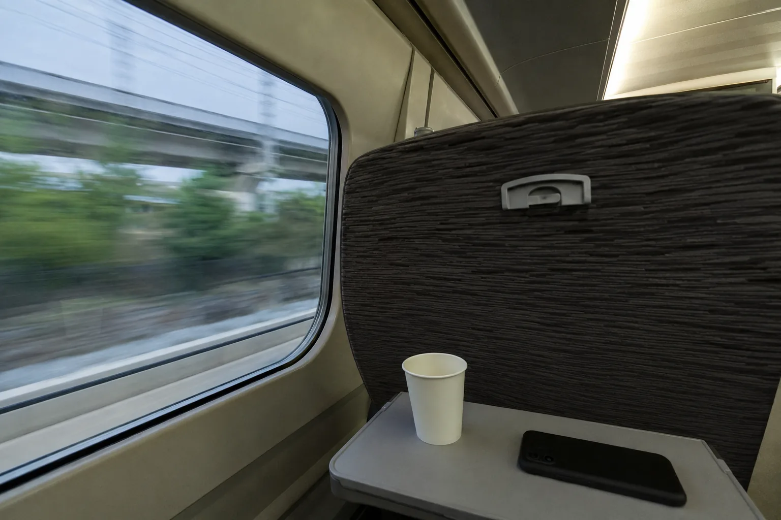The interior of a high-speed train from a seat: a folding tray table with a paper cup and a phone face-down, the seat back in front, and a blurred smear of green and concrete out of the window. Fluorescent light, banding in the ceiling panel.