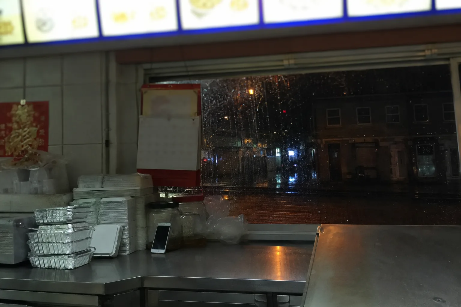 A takeaway counter photographed from behind the till at night: stainless steel prep surface, a stack of foil containers, a wall calendar, a phone propped against a jar, and the dark street through rain on the glass. Menu board out of focus. Nobody in shot.