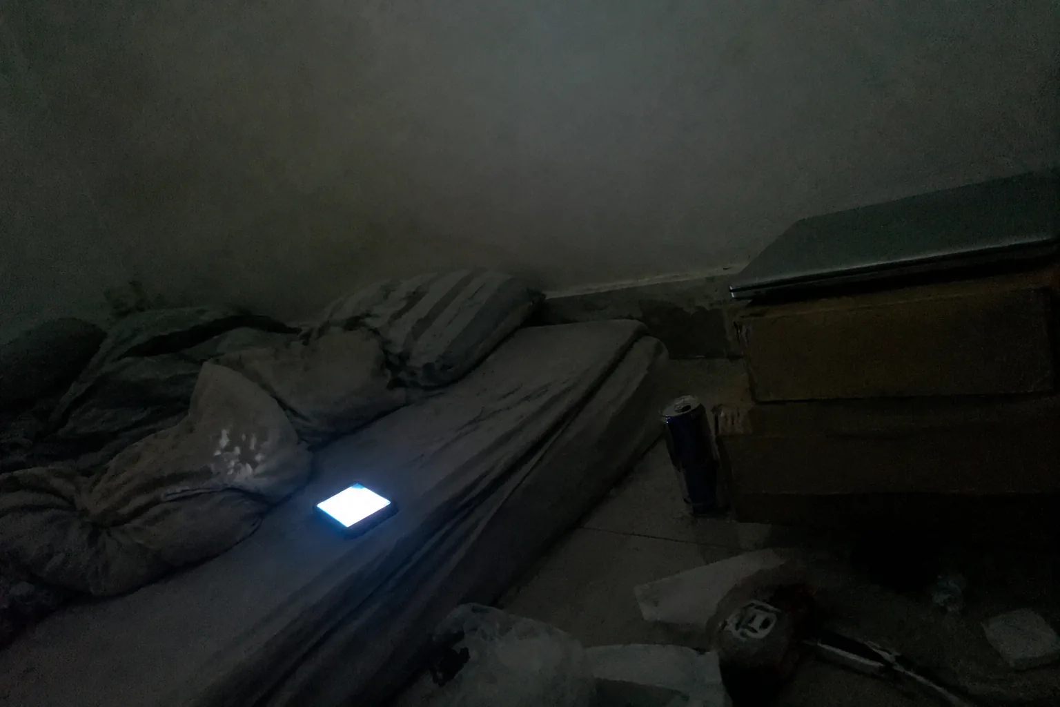 A very small rented room at night from floor level: a mattress on the floor with rumpled bedding, a closed laptop on a stack of boxes, an energy-drink can, and the cold blue-white of a phone screen face-up on the sheet as the only light. Heavy sensor noise.