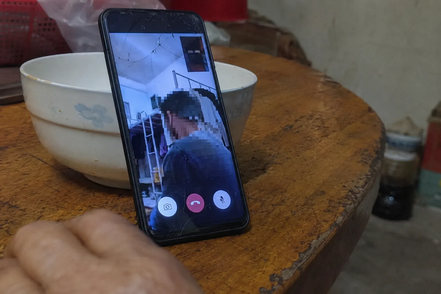 A video call on a cracked phone propped against a rice bowl on a worn kitchen table in a rural house. On the screen, a badly lit dormitory and a figure turned partly away with compression breaking their face into blocks. An older woman’s hand rests out of focus in the foreground.