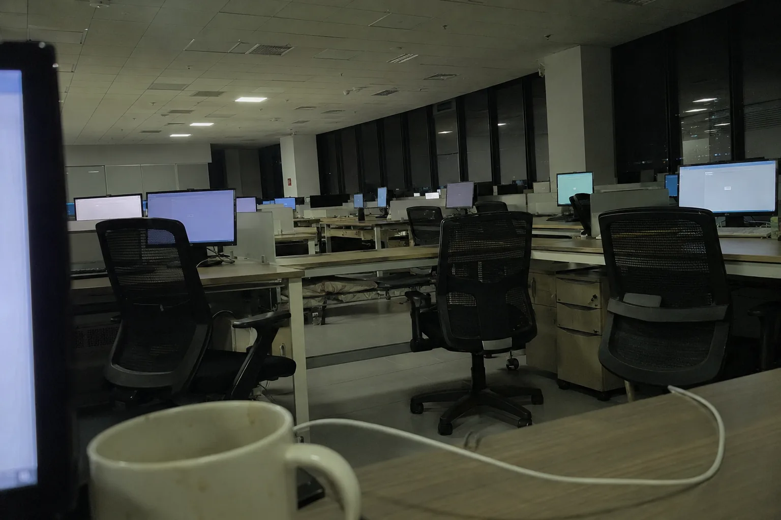An open-plan office at night from one desk: rows of monitors still on with nobody at them, ceiling fluorescents, a camp bed folded under a desk two rows away, a mug and a phone charger in the foreground. Green-grey cast.