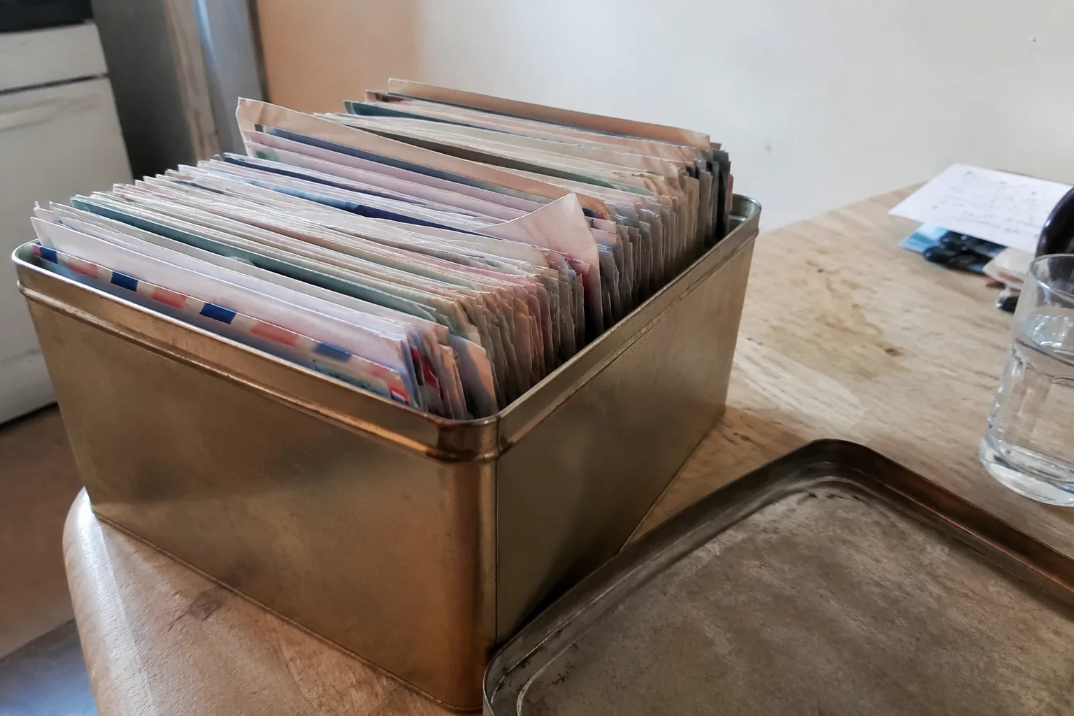 A biscuit tin open on a kitchen table in a British flat, packed upright with dozens of thin airmail envelopes with red-and-blue edge striping, the paper soft and handled. Grey window daylight on one side and a warm ceiling bulb on the other. The handwriting is out of focus.